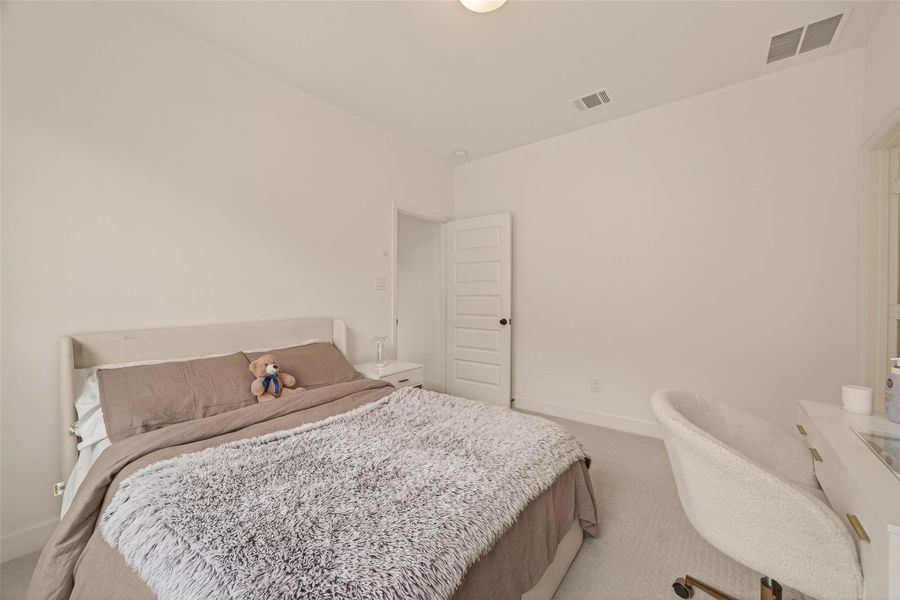 This is a cozy bedroom with neutral tones, featuring a bed with a plush comforter, a side table, and a desk with a chair. The room has a minimalist design with a soft carpet and a white door.