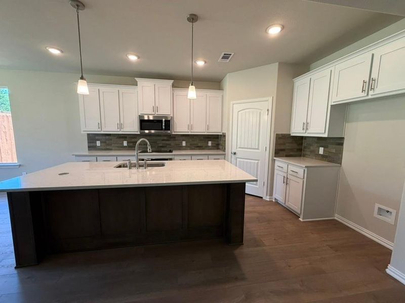 Modern kitchen featuring a large central island with a built-in sink and light-toned countertop, white cabinetry with brushed nickel hardware, a multi-toned tile backsplash, stainless steel microwave, and wood-finish flooring