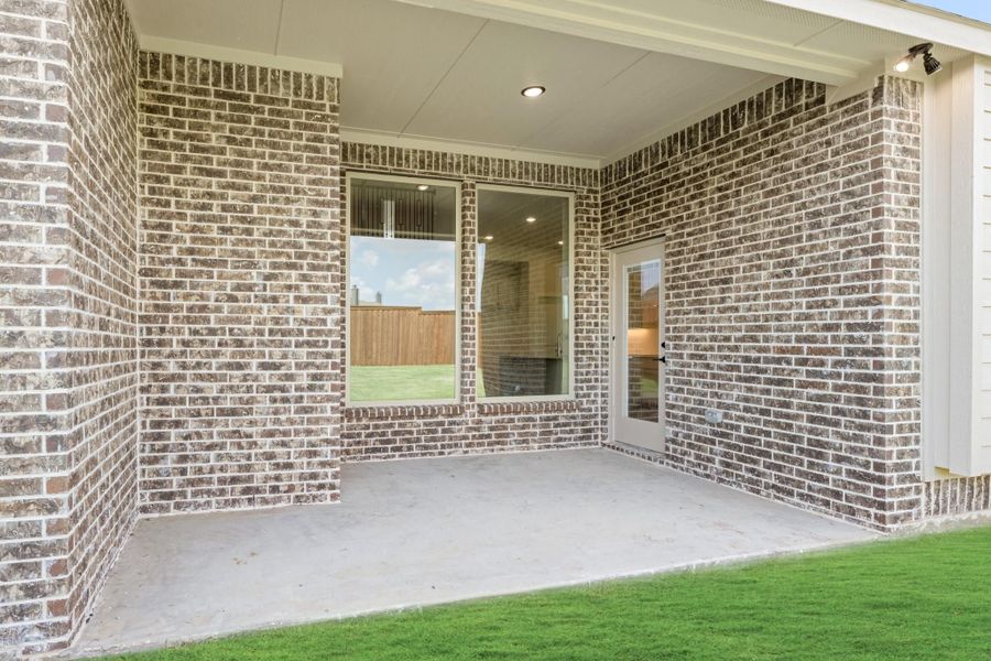 Covered back patio with concrete floor, brick exterior, large window, and glass door leading inside.