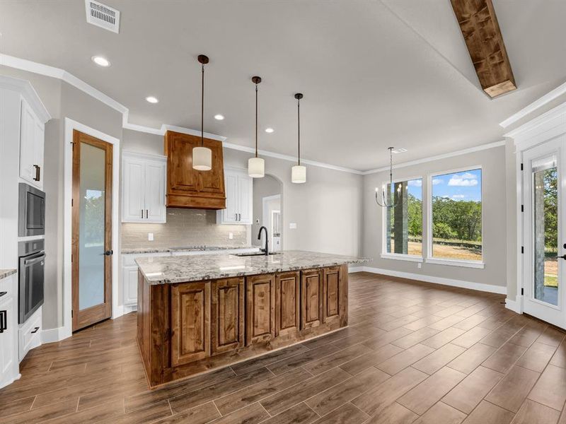 Kitchen with brown cabinets, pendant lighting, decorative backsplash, light stone counters, and a center island with sink