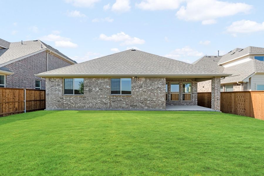 Rear view of single-story brick home with covered patio, large green lawn, and wood privacy fence