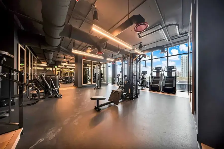 Fitness facility featuring industrial-style exposed ceiling ductwork, rubberized flooring, floor-to-ceiling windows, and integrated linear lighting fixtures Fitness facility featuring industrial-style exposed ceiling ductwork, rubberized flooring, floor-to-ceiling windows, and integrated linear lighting fixtures