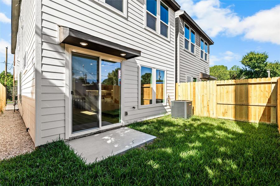 The private, fully fenced backyard features a manicured green lawn and a concrete patio for outdoor seating and grilling. Surrounded by a wooden privacy fence, the space includes a modern HVAC compressor and a covered awning with recessed lighting above massive sliding glass doors that lead inside.