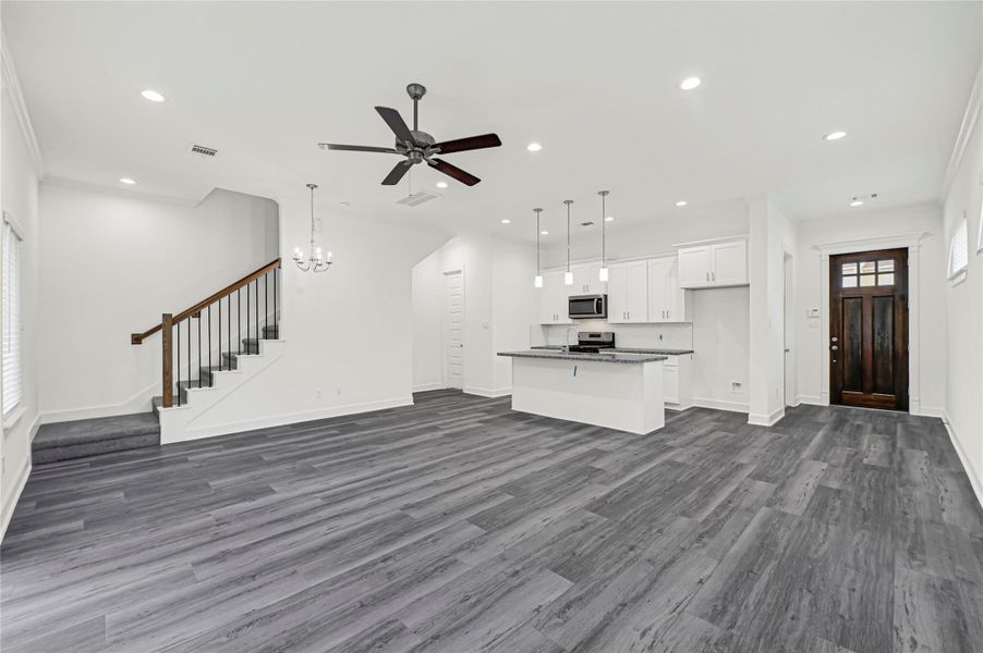 This photo showcases a modern open-concept living area with sleek gray flooring. It features a stylish kitchen with white cabinetry, stainless steel appliances, and a central island with pendant lighting. A dark wooden staircase leads upstairs, and a ceiling fan adds a touch of elegance. The space is bright and airy, ideal for comfortable living. This photo showcases a modern open-concept living area with sleek gray flooring. It features a stylish kitchen with white cabinetry, stainless steel appliances, and a central island with pendant lighting. A dark wooden staircase leads upstairs, and a ceiling fan adds a touch of elegance. The space is bright and airy, ideal for comfortable living.