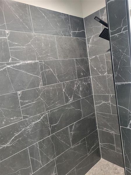 Shower stall featuring large format dark grey tile with white veining, a matte black rain shower head, and a complementary stone-finish shower pan