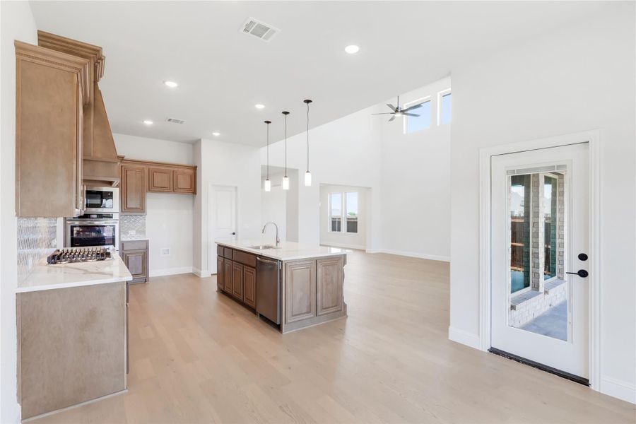 Open kitchen with wood cabinets, center island, pendant lights, and light hardwood floors in a high-ceiling space. Open kitchen with wood cabinets, center island, pendant lights, and light hardwood floors in a high-ceiling space.