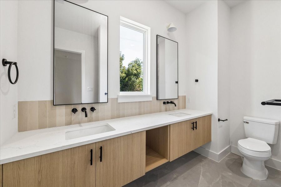 Warm wood cabinetry, quartz countertops, dual sinks, matte black fixtures, and full height tile continue the home’s cohesive design palette.