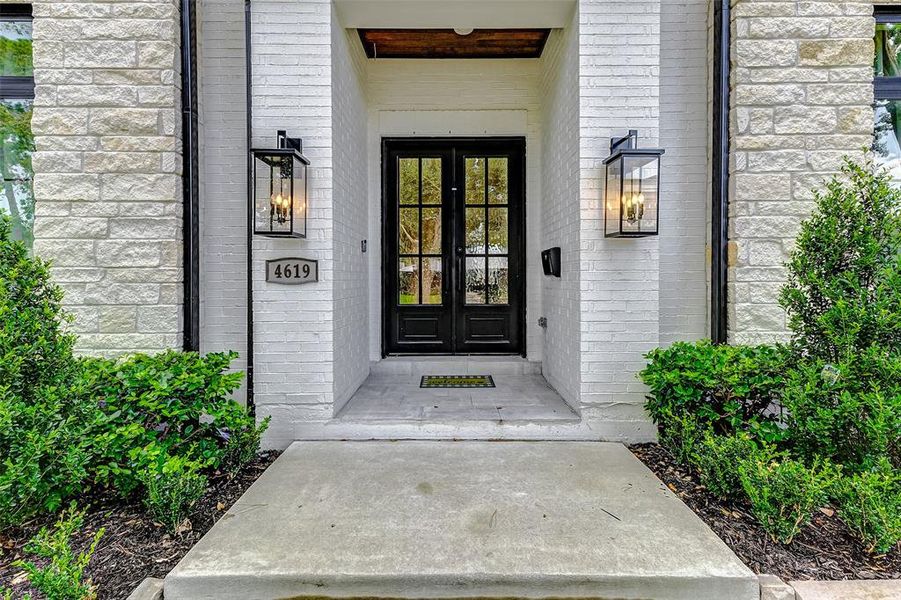 This elegant entryway features a modern black custom iron double glass front door framed by large glass panels, set against a white brick facade. Flanked by stylish lanterns and lush greenery, it offers a welcoming and sophisticated first impression.