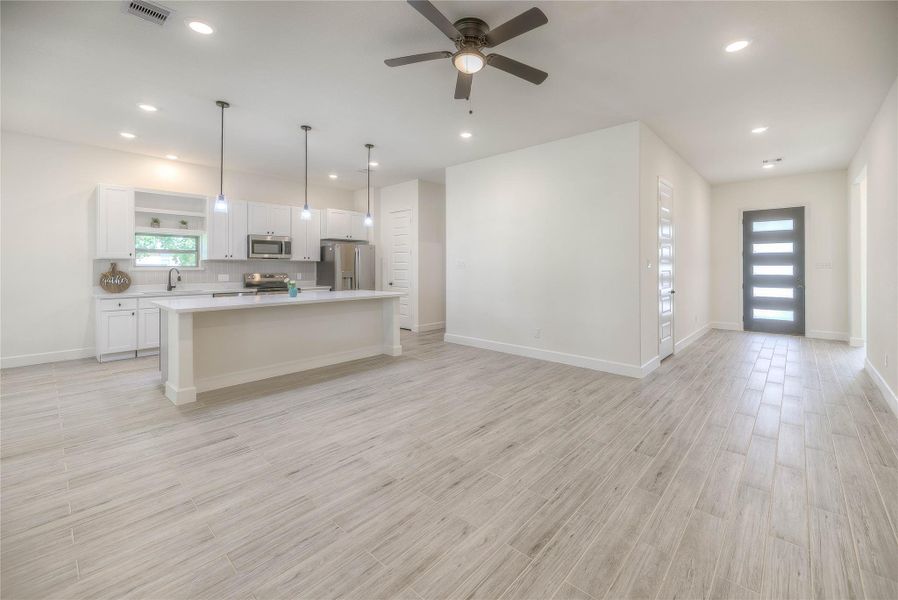 Spacious, open-concept living area with light wood flooring, modern recessed lighting, and a ceiling fan. The kitchen features a large island, white cabinetry, stainless steel appliances, and pendant lights, creating a bright and inviting space. View to the the front door.