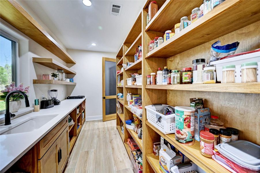 This photo shows a spacious, well-organized pantry with ample wooden shelving and storage space. It features a countertop with a sink, open shelves for easy access, and natural light from a window, creating a bright and functional space.