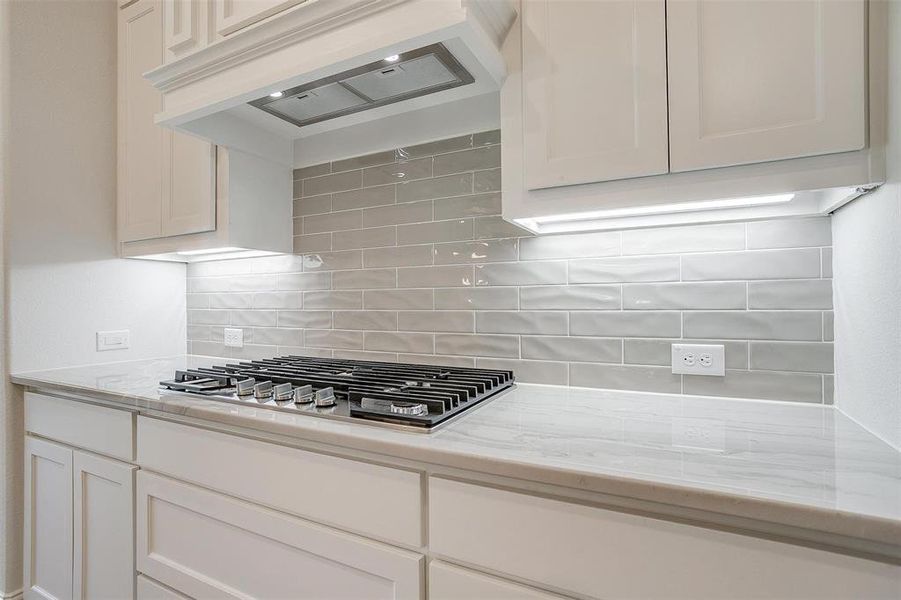 Kitchen featuring stainless steel gas cooktop, premium range hood, light stone countertops, and decorative backsplash