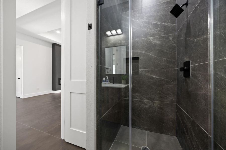 This photo showcases a modern shower with sleek, dark tiles and a glass enclosure. The adjacent room features a clean, minimalist design with large tiles and white walls, offering a contemporary aesthetic.