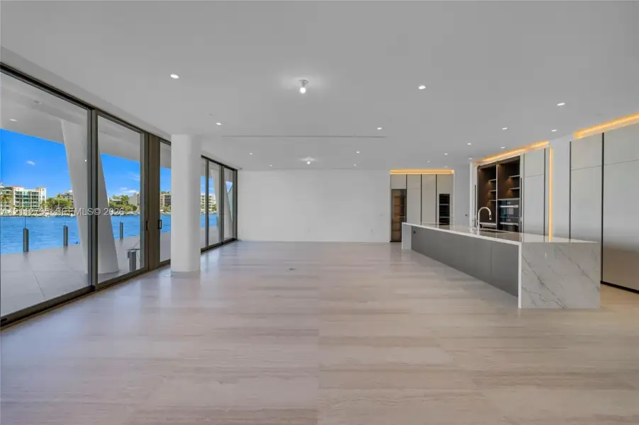 Open-concept interior featuring light-toned stone-finish flooring, floor-to-ceiling glass sliding doors, recessed lighting, and a kitchen island with a stone-finish countertop and integrated cabinetry