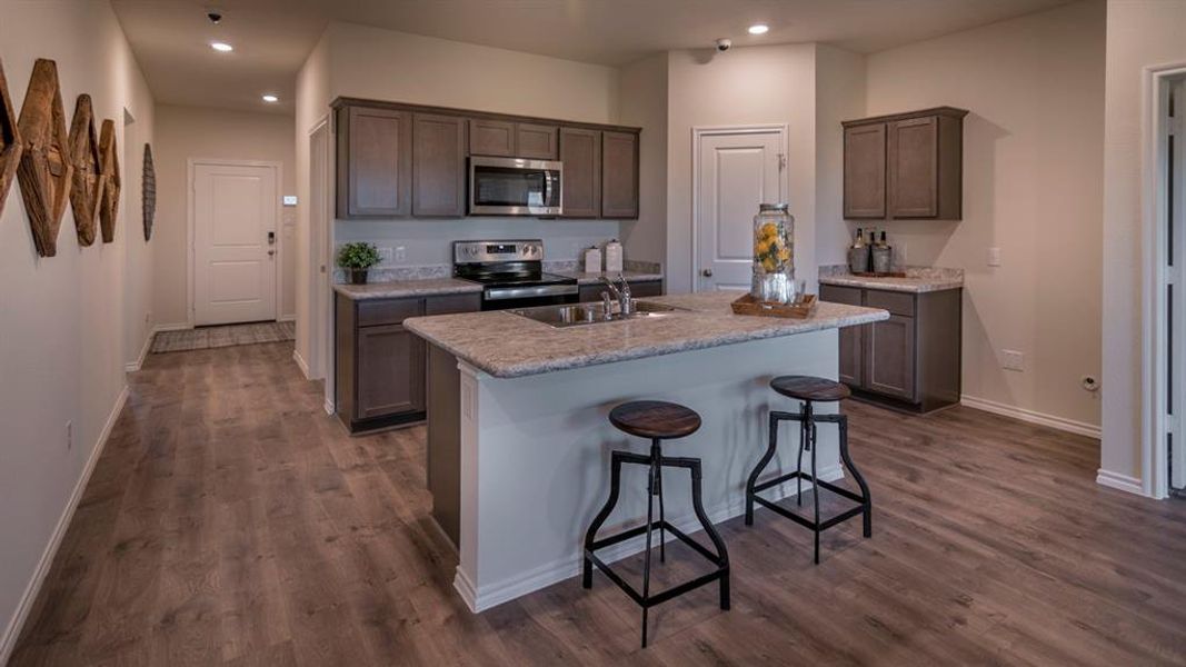 Open-concept kitchen featuring an island with a sink, stainless steel appliances, dark wood-finish cabinetry, and light-colored countertops
