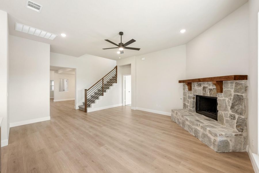 Open living room with stone fireplace, wood mantel, hardwood floors, ceiling fan, and staircase with metal railing