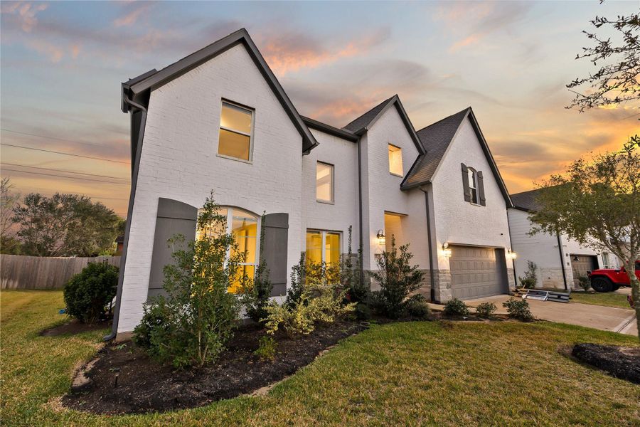 This charming two-story home features a white brick exterior with dark trim, a spacious front yard with manicured landscaping, and a two-car garage. The warm lighting from the windows complements the serene sunset backdrop. This charming two-story home features a white brick exterior with dark trim, a spacious front yard with manicured landscaping, and a two-car garage. The warm lighting from the windows complements the serene sunset backdrop.