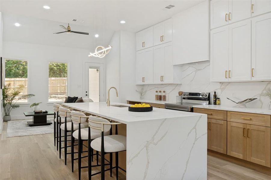 Spacious kitchen featuring a large waterfall island with integrated sink, white shaker cabinetry, light wood-finish lower cabinetry, a light-toned backsplash, and stainless steel appliances Spacious kitchen featuring a large waterfall island with integrated sink, white shaker cabinetry, light wood-finish lower cabinetry, a light-toned backsplash, and stainless steel appliances