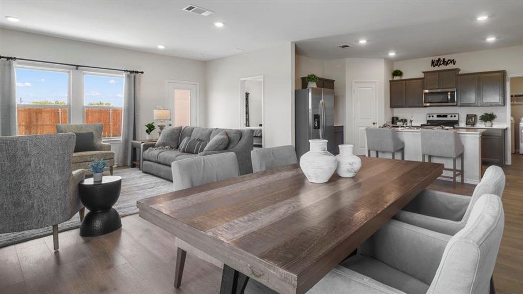 Spacious open-concept interior featuring wood-finish flooring, recessed lighting, a modern kitchen with dark cabinetry and stainless steel appliances, and a dining area with a rustic wood table
