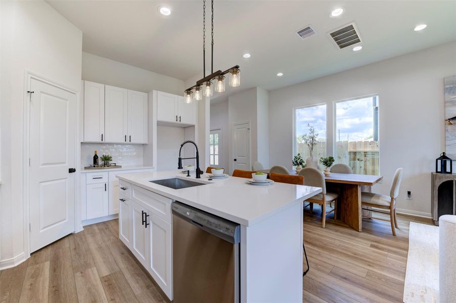 Kitchen island with quartz countertop, farmhouse sink, matte black coil-spring faucet, stainless dishwasher, and Edison rail pendant overhead. Pantry door and appliance niche visible on left. Open sightline to dining area and large rear windows with backyard view.