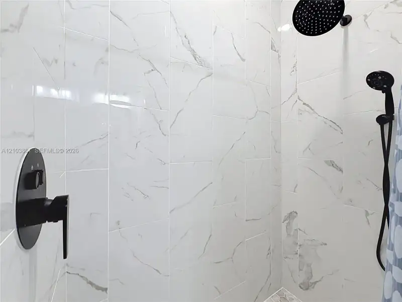 Walk-in shower featuring large-format stone-patterned wall tile, matte black rainfall showerhead, adjustable handheld shower wand, and a matching matte black pressure-balance faucet