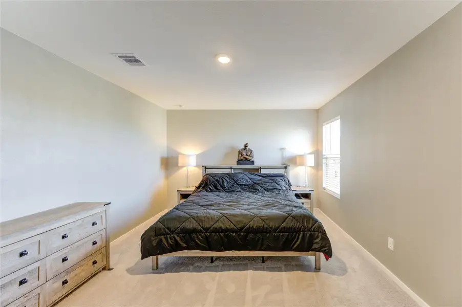 This spacious bedroom features neutral walls and carpeting, a comfortable bed with side tables and lamps, and a large window providing natural light. A dresser offers additional storage.