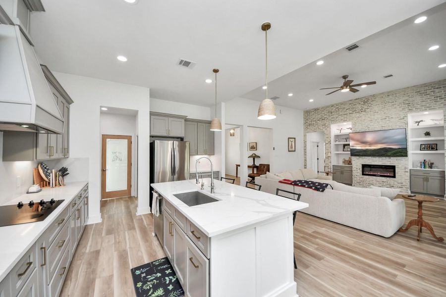 Experience open-concept living with a modern kitchen featuring sleek cabinetry and a central island, flowing seamlessly into a cozy living area with a stylish brick accent wall and built-in fireplace. Experience open-concept living with a modern kitchen featuring sleek cabinetry and a central island, flowing seamlessly into a cozy living area with a stylish brick accent wall and built-in fireplace.