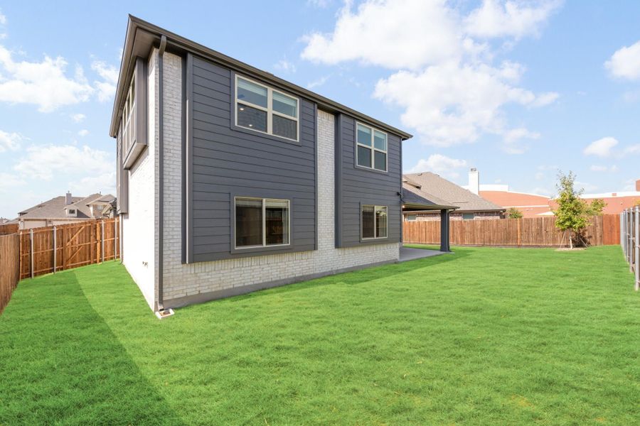 Two-story home with gray siding, white brick base, covered patio, and fenced backyard with green lawn