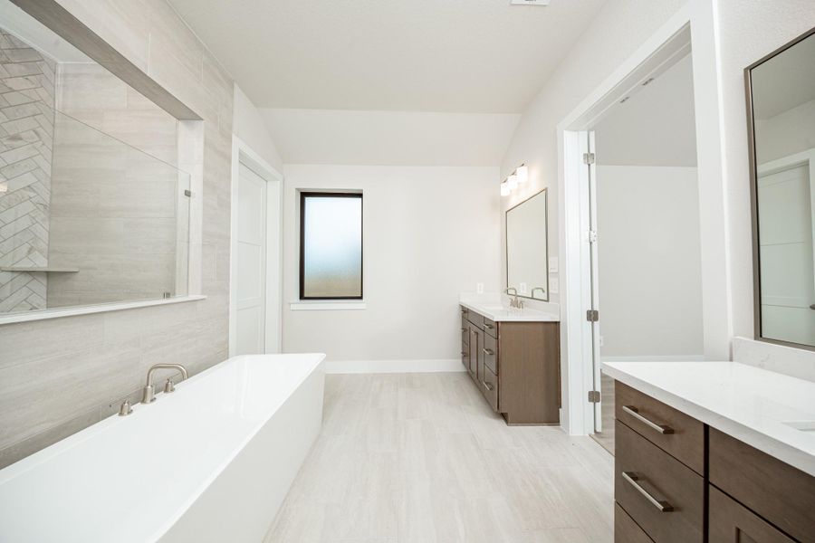 This spa-inspired primary bathroom showcases a sleek freestanding soaking tub set against a striking tiled accent wall with a glass-enclosed shower beyond. Tile flooring, clean modern lines, and brushed nickel fixtures create a serene and contemporary retreat filled with natural light.