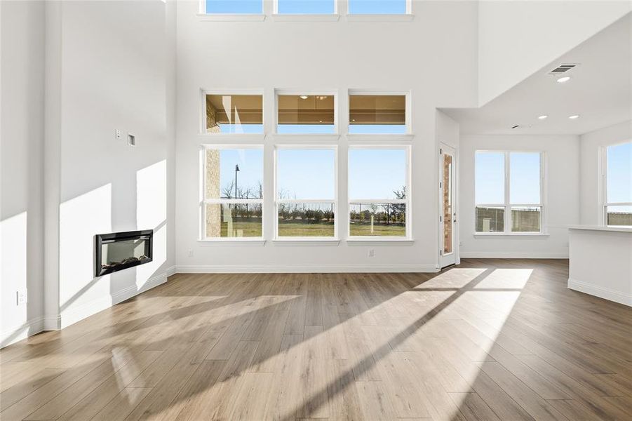 Unfurnished living room featuring a glass covered fireplace, healthy amount of natural light, light wood-style flooring, and a high ceiling Unfurnished living room featuring a glass covered fireplace, healthy amount of natural light, light wood-style flooring, and a high ceiling