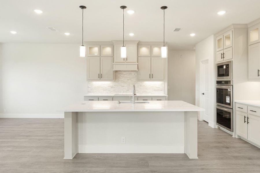 White kitchen with large island, pendant lights, stone backsplash, and double wall ovens