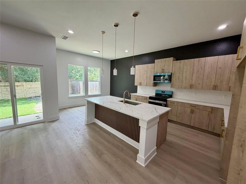 Open-concept kitchen featuring an island with a white countertop and integrated sink, light wood-finish cabinetry, and a black wall accent Open-concept kitchen featuring an island with a white countertop and integrated sink, light wood-finish cabinetry, and a black wall accent