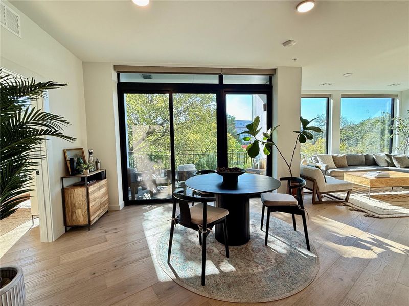 Natural light floods this dining area, featuring sleek wood flooring and a view of a tranquil balcony.