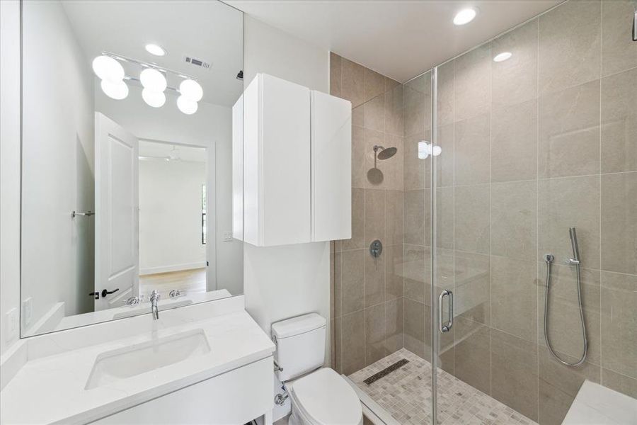 Secondary Bedroom No. 1’s ensuite bathroom features a walk-in shower with designer grey tile, a frameless glass door, and chrome plumbing fixtures. The space is enhanced by quartz countertops, designer porcelain floor tile, and a custom vanity with chrome cabinet hardware. A wall-mounted designer chrome faucet completes the luxurious look.