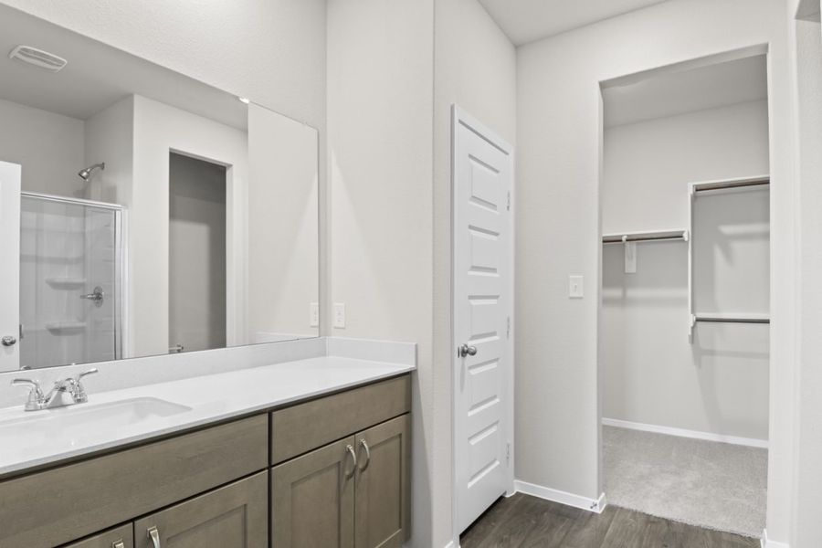 Image of a primary bathroom with brown cabinets, a white double vanity, a large mirror and a connecting walk-in closet