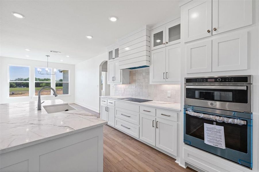 Contemporary kitchen featuring bright white cabinetry, a large center island with an undermount sink, built-in stainless steel appliances, wood-finish flooring, and a tile backsplash