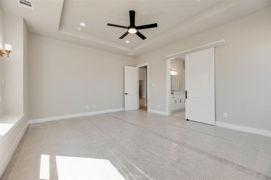 Unfurnished bedroom with light carpet, a ceiling fan, recessed lighting, a tray ceiling, and connected bathroom