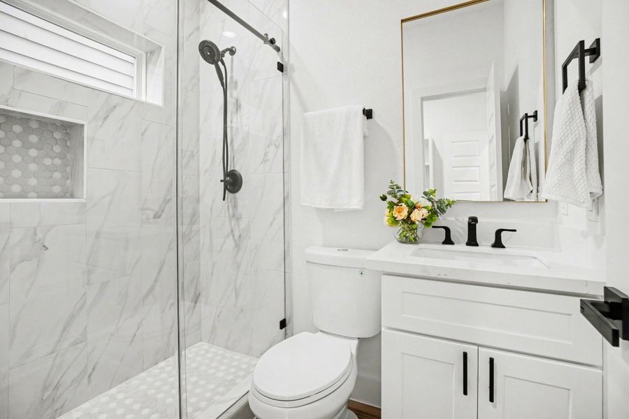 This bathroom features a modern design with a glass-enclosed shower, marble-style tiles, and a built-in niche. It includes a sleek vanity with black fixtures and a large mirror, offering a clean and elegant look.