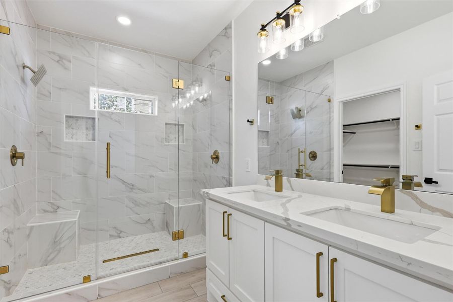 Primary bathroom featuring double vanity with gold fixtures, frameless glass walk-in shower, and marble look tile.