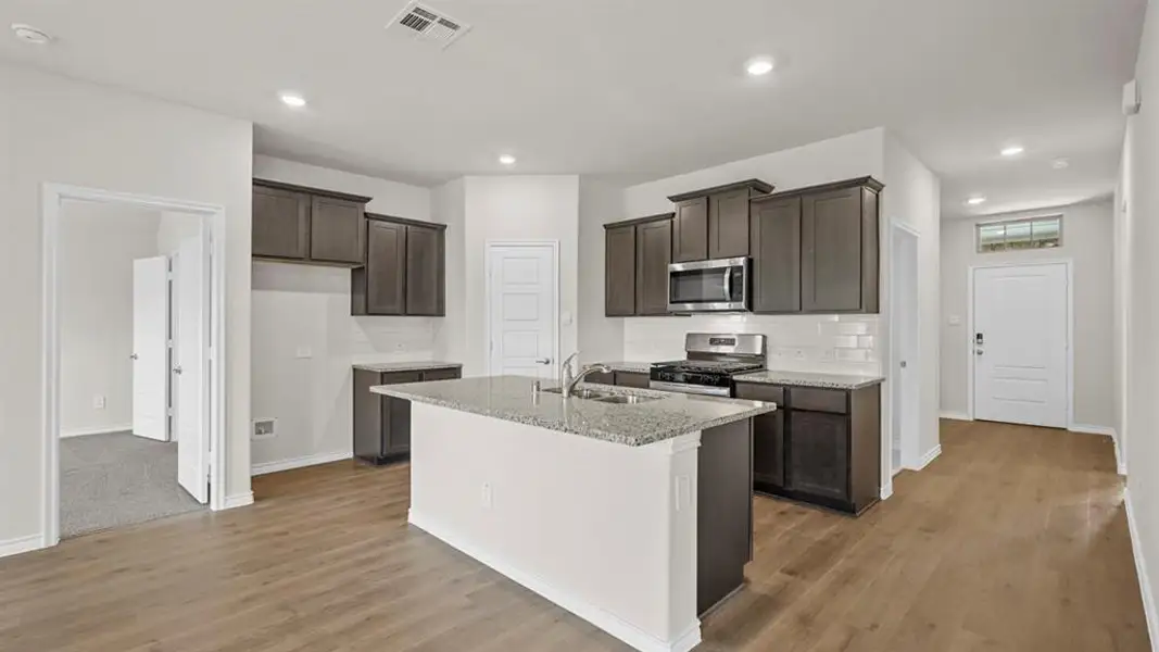 Open-concept kitchen featuring an island with a sink, granite countertops, wood-finish flooring, stainless steel appliances, and dark wood cabinetry