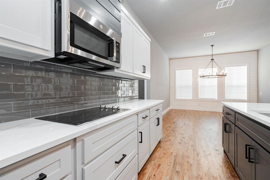 Kitchen with black electric stovetop, a chandelier, stainless steel microwave, decorative backsplash, and visible vents Kitchen with black electric stovetop, a chandelier, stainless steel microwave, decorative backsplash, and visible vents