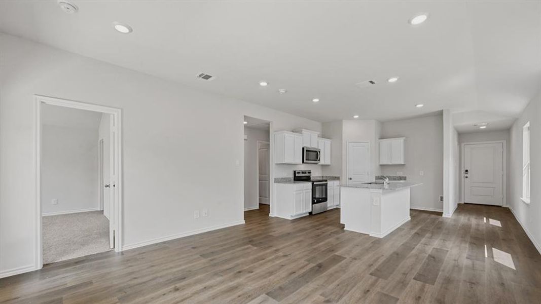Open-concept living area featuring wood-finish flooring, recessed lighting, and a kitchen with white cabinetry, stainless steel appliances, and an island with a light-toned countertop