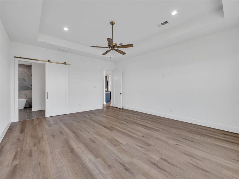 Empty room featuring light hardwood / wood-style flooring, a barn door, a tray ceiling, and ceiling fan