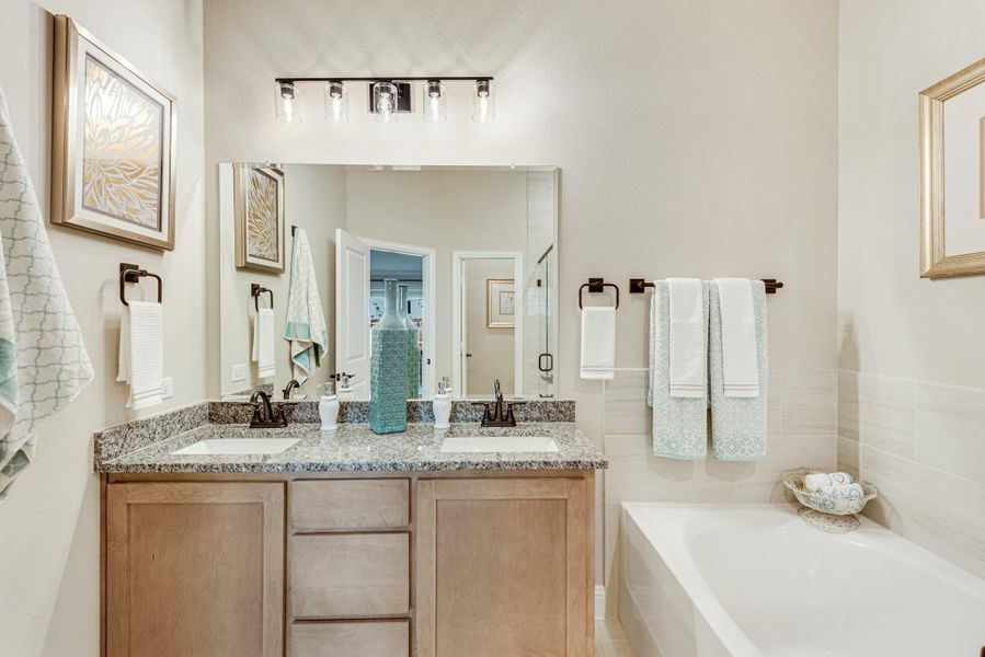 Bathroom with double sink vanity, granite countertop, large mirror, and soaking tub beside tiled wall.