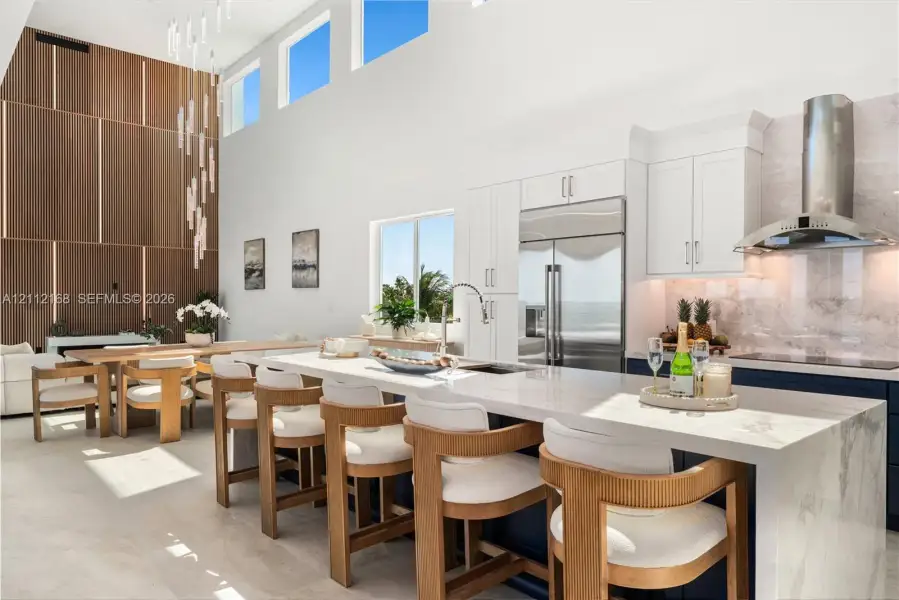 Kitchen featuring a waterfall island with stone countertop, cabinetry with integrated appliances, vertical wood-paneling wall feature, soaring ceilings with transom windows, and a multi-pendant light fixture Kitchen featuring a waterfall island with stone countertop, cabinetry with integrated appliances, vertical wood-paneling wall feature, soaring ceilings with transom windows, and a multi-pendant light fixture