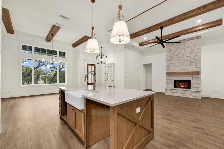 Kitchen featuring brown cabinets, beam ceiling, hanging light fixtures, a center island with sink, and a fireplace Kitchen featuring brown cabinets, beam ceiling, hanging light fixtures, a center island with sink, and a fireplace