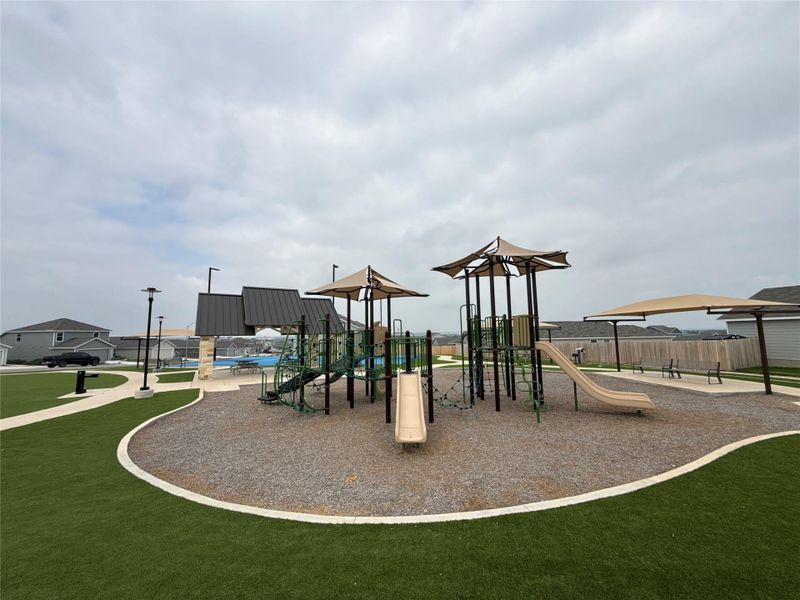 Outdoor recreation area featuring a playground with multiple slides and climbing structures, shaded seating areas, and expansive artificial turf