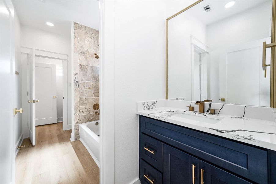 Adjoining bath featuring a beautifully tiled tub/shower surround and an ash-blue vanity that continues the home’s cohesive color palette, paired with quartz counters and brushed gold accents for a polished finish.