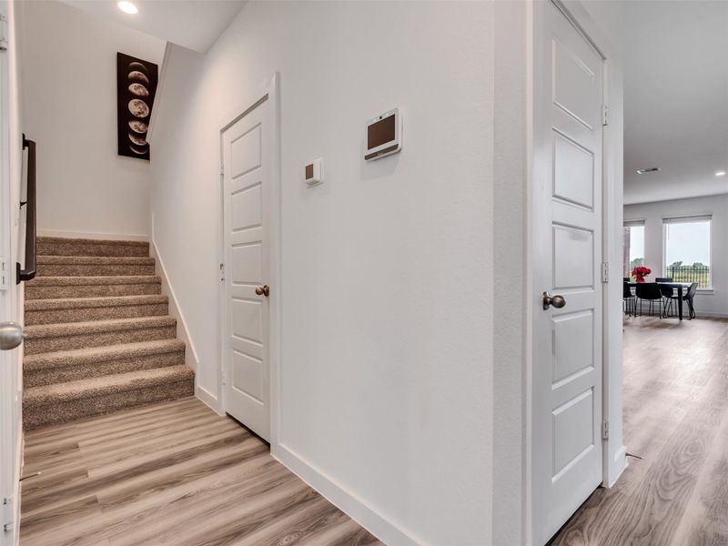 This photo showcases a modern entryway with light wood flooring, carpeted stairs, and a view into a bright dining area with large windows. The walls are painted white, creating a clean and inviting atmosphere.