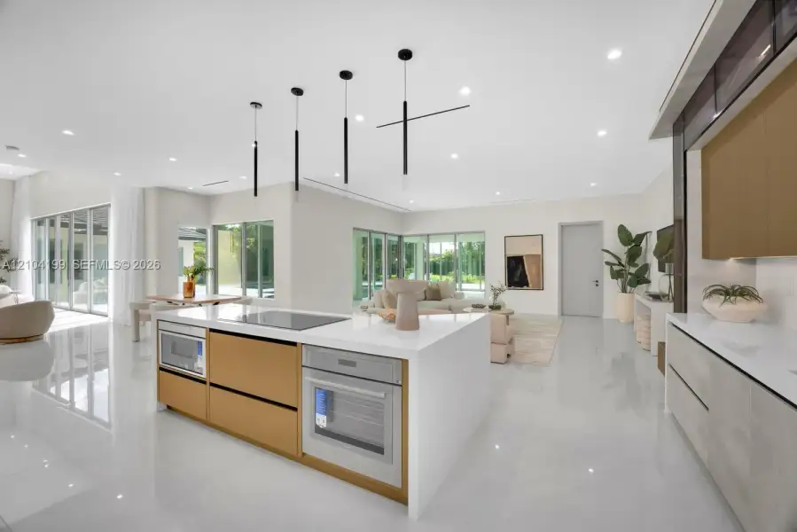 Modern kitchen featuring a central island with integrated cooktop and oven, sleek flat-panel cabinetry, light-toned stone-surface countertops, recessed ceiling lighting, and minimalist linear pendant fixtures
