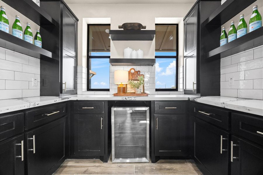 Kitchen with open shelves, dark cabinetry, beverage cooler, light stone countertops, and tasteful backsplash Kitchen with open shelves, dark cabinetry, beverage cooler, light stone countertops, and tasteful backsplash